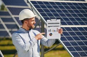 Pointing to the notepad with,, diagrams ,and graphs. Engineer with photovoltaic solar panels outdoors at daytime photo