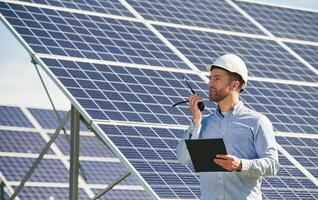 Using walkie talkie ,and,, holding notepad. Engineer with photovoltaic solar panels outdoors at daytime photo