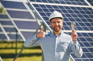 Smiling, and holding bulb. Engineer, with photovoltaic solar panels outdoors at daytime photo