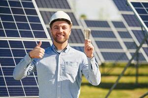Smiling, and holding bulb. Engineer ,with, photovoltaic solar panels outdoors at daytime photo