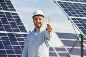 Smiling and holding bulb. Engineer with ,,photovoltaic solar panels outdoors at daytime photo