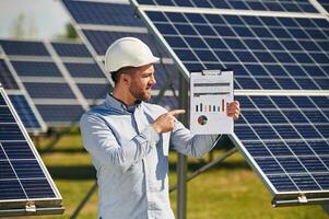 Pointing, to the notepad with diagrams and graphs. Engineer with photovoltaic solar panels outdoors at daytime photo