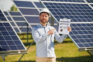 Pointing to, the notepad with diagrams and graphs. Engineer with photovoltaic solar panels outdoors at daytime photo