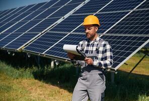 On, th,e field with notepad in hands. Engineer with photovoltaic solar panels outdoors at daytime photo