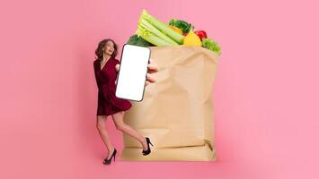 A customer joyfully holds a smartphone while standing beside a large paper bag filled with fresh groceries. The backdrop features a bright pink color, highlighting the new shopping trend. photo