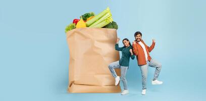 Two happy shoppers stand beside a large paper bag filled with fresh vegetables and fruits. They express joy and enthusiasm about their grocery purchase, contributing to the new normal of shopping. photo