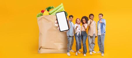 Five individuals smile while showcasing a smartphone in front of a paper grocery bag filled with fresh produce. This reflects modern shopping trends in an isolated background. photo