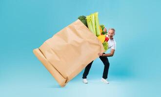 A customer holds a large paper bag overflowing with fresh vegetables and fruits while standing against a vibrant blue backdrop. This reflects a modern shopping experience during changing times. photo