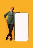 Mobile Offer. Happy Black Guy Leaning At Big Smartphone With Blank White Screen, Cheerful African Man Demonstrating Copy Space For Your Design Or Advertisement, Posing Over Yellow Background, Mockup photo