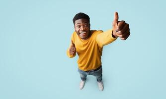 Top view of joyful handsome african american millennial guy in nice outfit showing thumb up and smiling at camera on blue studio background, recommending something exciting, copy space, full length photo