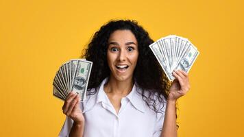 Surprised woman holding lots of money in both hands and exclaiming with excitement, lucky winner lady standing with bunch of dollars over yellow studio background, looking at camera, copy space photo