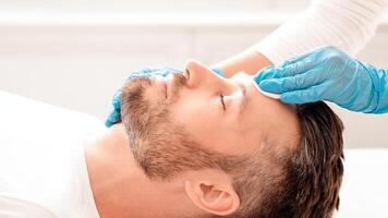 Side view of unrecognizable cosmetologist cleansing bearded man face with toner before or after treatment, using cotton pad. Middle-aged businessman having beauty procedures at spa photo