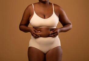 Body Neutrality Concept. Closeup Cropped View Of Unrecognizable Chubby African American Woman In Underwear Holding Fat Fold Love Handles On Her Belly, Standing Isolated Over Brown Studio Background photo