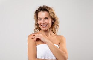 Portrait Of Pretty Middle Aged Female Wrapped In Bath Towel Posing Over Light Grey Background, Attractive Woman With Beautiful Skin Smiling At Camera, Enjoying Self-Care Treatments, Copy Space photo