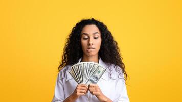Portrait Of Thoughtful Young Woman With Bunch Of Dollar Banknotes In Hands Posing Over Yellow Background In Studio, Thinking How To Spend Money Wisely, Biting Lip With Excitement, Copy Space photo