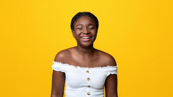 Portrait of beautiful african american lady sincerely laughing on yellow background, positive black woman having good mood, emotionally reacting to joke, joyful female smiling with closed eyes photo