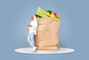A customer, wearing casual attire and a hijab, holds a smartphone while leaning against a large paper bag filled with fresh vegetables and fruits. photo