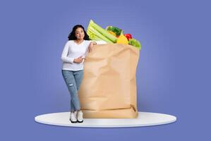 A shopper stands next to a large paper bag filled with fresh fruits and vegetables. The background is a simple blue, emphasizing the joy of purchasing groceries in the new normal. photo