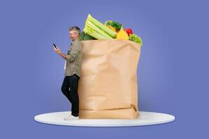 A customer stands beside a large paper bag filled with fresh groceries. He checks his phone, adapting to modern shopping trends at home, showcasing a blend of convenience and isolation. photo