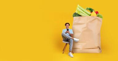 A young person sits casually on a chair, smiling while using a mobile device. Behind them is a large paper bag filled with fresh vegetables, highlighting the convenience of grocery shopping at home. photo