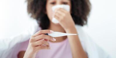 First symptoms of illness, fever, flu, colds and covid virus. Sad blurred millennial african american woman in blanket wipes nose with napkin and looks at thermometer, selective focus, free space photo