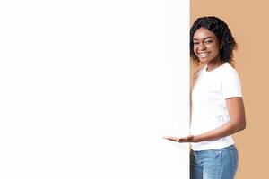 Attractive black woman showing blank advertisement board, standing on yellow studio background. Smiling african american lady posing next to big white placard for text or advertising, panorama photo