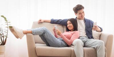 Korean Married Couple With Smartphones Browsing Internet And Using Apps Relaxing Sitting On Couch In Living Room At Home. Gadget Lifestyle, Mobile App Concept. Free Space For Text photo