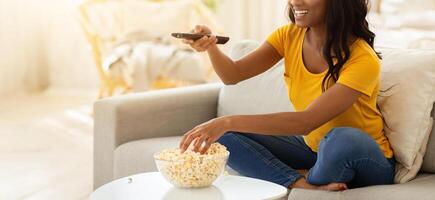 Young African American woman sitting on couch, eating popcorn and watching interesting show on television, indoors. Pretty black lady with TV remote switching channels at home photo