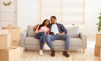 Cheerful millennial family moving to new property, taking break from unpacking, resting on sofa among carton boxes. African American family renting or purchasing new house, hugging on couch photo