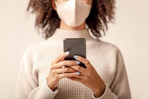 News about spread of virus in world and social networks. African americans lady in protective mask and sweater typing on smartphone isolated on light cream background, free space, studio shot photo