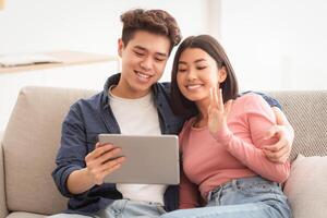 Asian Couple With Digital Tablet Making Call Online Waving Hand To Webcam Sitting On Sofa At Home. Chinese Family Calling Via Computer. Modern Remote Communication Concept photo