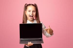 Wow, look there. Happy little girl with open mouth shows at laptop with blank screen, isolated on pink background, copy space photo