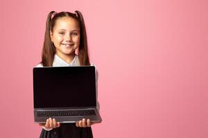 Remote education during quarantine. Smiling girl in school uniform holding laptop with blank screen, isolated on pink background photo