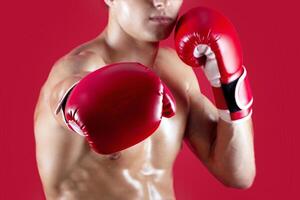 Determined young boxer in fighting gloves making punch at camera on red studio background, selective focus. Unrecognizable millennial sportsman with bare chest fighting, cropped view photo