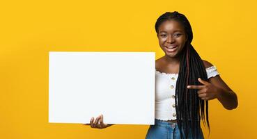 Cheerful Black Lady Holding And Pointing At White Blank Placard, Demonstrating Free Place For Your Text Or Advertisement, Showing Copy Space On Billboard, Posing Over Yellow Studio Background photo