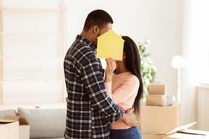 Young black couple hiding behind paper house and hugging while relocating to their new apartment. African American boyfriend and girlfriend moving into recently purchased home photo