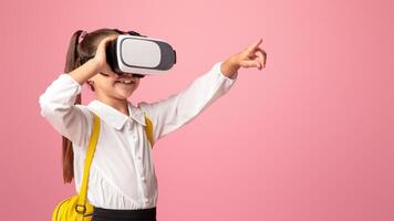 Shocked schoolgirl with virtual reality glasses pointing finger aside on pink background in studio on pink background, studio shoot photo