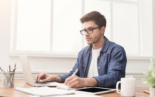 Young concentrated man typing on laptop and taking notes, in modern office. Handsome programmer working on computer and writing message, copy space photo