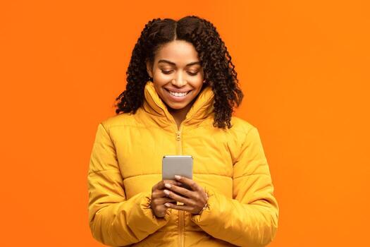 Cheerful Black Lady With Cellphone Using Mobile App, Texting And Browsing Internet Standing Over Yellow Background In Studio, Wearing Winter Clothes. Smartphone User Using Cellphone App Concept photo