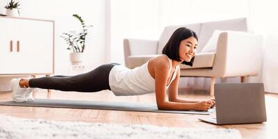 Training At Home. Full length portrait of sporty smiling asian woman doing plank while watching online tutorial on laptop computer, exercising in living room on the floor on yoga mat, free space photo