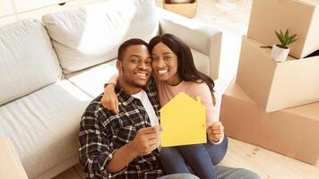 Black millennial guy with his girlfriend hugging and holding paper house while sitting on floor of their new apartment on moving day. Relocation, tenancy, home rentals concept photo