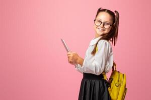 Child using gadgets to study. Cheerful schoolgirl in glasses with backpack holds digital tablet and looks at camera, on pink background, copy space photo