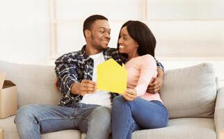 Black millennial woman with her boyfriend hugging and holding paper house in their new apartment, selective focus. Positive young homeowners resting on couch during relocation, panorama photo