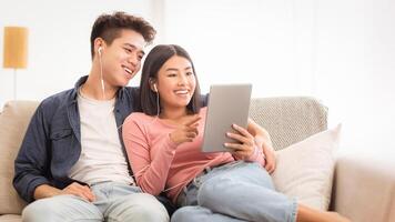 Cheerful Asian Couple Using Digital Tablet Browsing Internet Together Sitting Embracing On Couch At Home On Weekend. Korean Family Enjoying Online Movie On Computer, Gadgets And Apps. photo