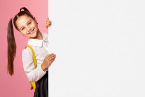 Ready for lesson. Cheerful schoolgirl in uniform peeps out from with white billboard with copy space, studio shoot photo