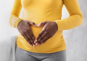 Pregnant black woman holding palms in heart shape on her big tummy, cropped. Unrecognizable african american expecting lady standing by window at home. Pregnancy and maternity concept photo