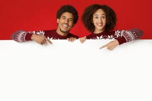 Happy New Year and winter holidays celebration. Smiling millennial african american lady and man in same Xmas sweaters holds white billboard and points at it with fingers, isolated on red background photo