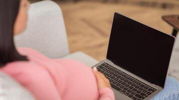 Pregnant Lady Using Laptop With Empty Screen Sitting On Sofa At Home. Pregnancy Blog. Unrecognizable Asian Woman Browsing Internet On Computer Waiting For Baby. Mockup, Cropped, Selective Focus photo