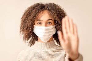 Stop coronavirus and health protection during epidemic. Serious african american young female in protective mask making hand gesture, focus on face of lady in sweater isolated on light background photo