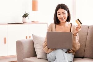 Online Payment Concept. Smiling asian woman making purchases via internet, using computer, holding and showing credit card. Satisfied lady with laptop sitting on couch, free space photo